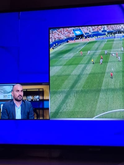 This is a photograph taken of a television screen displaying a football match. On the left side of the screen, a man in a dark suit and light blue shirt is seated, looking towards the right. He has a shaved head and a beard. Behind him, there appears to be a shelf with some decorative items. The right side of the screen shows a football field with several players in red and white uniforms. The field is green with white lines, and a crowd can be seen in the background. The television screen itself is placed against a bright blue background. There is no information in the image to suggest a location in Nova Zagora, Bulgaria.