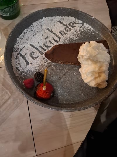 A slice of chocolate cake is presented on a dark speckled plate, dusted with powdered sugar that spells out "Felicidades." To the right of the cake is a dollop of whipped cream. To the left of the cake are a raspberry, a blackberry, and a strawberry with a candle inserted. The plate is on a wooden surface. A green bottle is partially visible on the left side of the image. The scene appears to be in a restaurant or dining setting in Huixquilucan de Degollado, Mexico.