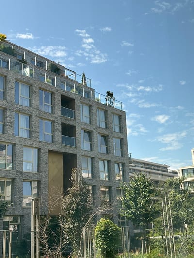 The image depicts a modern apartment building constructed with gray bricks, featuring numerous large windows and several balconies with glass railings. The building has a stepped facade on the upper floors, with some balconies featuring greenery. The sky above is bright blue with scattered white clouds. In the foreground, there are trees and vegetation, including some bare branches and lush green bushes. In the background, other buildings are visible, including a long, multi-story structure with balconies and a lighter-colored facade. This building is located at Houthaven, Amsterdam, Netherlands.