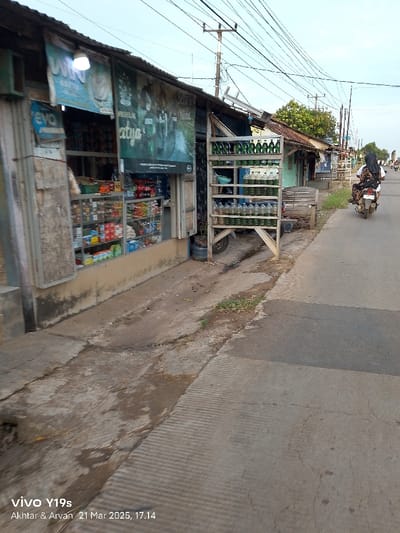 The scene is a narrow, paved alleyway in Anjatan, Indonesia. The alleyway is lined with small, simple buildings, one of which is a shop with a glass front. The shop has various products displayed on shelves inside, including bottles of drinks, packaged food, and other miscellaneous items. There is an advertisement for a motorcycle brand on the side of the shop, next to a sign with the words "Saya" and "Atyo" printed on it. There are also many power lines running across the alleyway. A metal rack full of green and clear plastic bottles stands in front of the shop. There is a person riding a motorcycle in the distance, and some plants are growing in the cracks of the pavement. The alleyway is empty except for the rider and the motorcycle. The time is approximately 5:14 PM.
