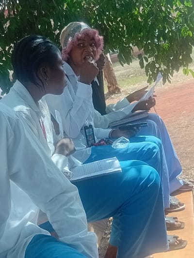 Several people sit outdoors beneath the shade of a tree. They are dressed in white shirts and blue pants or skirts. Some have notebooks and papers in their hands, while others hold cell phones. One person has pink hair and another wears a patterned cap. The group seems to be focused on reading or writing, possibly in a class or meeting. Some sandals are visible at the edge of the frame.