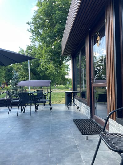 The image shows an outdoor seating area of a cafe. The setting is a paved patio with large grey tiles, adjacent to a building with wooden exterior panels and large glass windows. Several black metal tables and chairs are arranged on the patio. One table has a light purple umbrella partially covering it. In the foreground, a black umbrella stands open. Beyond the patio, a grassy area with trees is visible. The sky is bright and appears to be daytime, with some clouds.

Visible text on the window includes what appears to be business hours: "Mon-Fri 9:00-20:00", "Sat-Sun 10:00-21:00". There are also cartoonish drawings on the window, including figures that resemble Snoopy, and speech bubbles with "coffee" and "hi". The cafe's name may also be visible, though it is partially obscured. The scene suggests a pleasant day for outdoor dining or relaxing at a cafe. The location context is Znamianka, Ukraine.