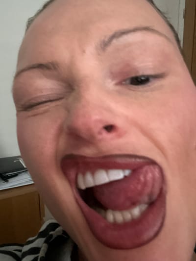 A close-up, front-facing image depicts an individual, likely female, in an indoor setting located in Wealden, United Kingdom. The subject's face fills the frame, presenting a playful expression: her left eye is closed in a wink, while her right eye remains open. Her mouth is wide open, exposing white teeth and a protruded tongue. The lips are colored with a dark red or burgundy lipstick. The skin appears light-toned, with minor facial creases around the winking eye. The background is largely out of focus, revealing a light-colored wall on the upper left, an indistinct dark object resting on papers atop a light brown wooden surface in the lower left, and a partially visible black and white striped garment at the bottom of the frame.