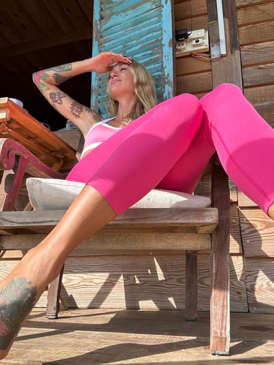 A woman with blonde hair and tattoos on her arms and legs is sitting on a wooden chair outside a building. She is wearing a pink sports bra and bright pink leggings. The woman is shielding her eyes from the sun with her hand. The background shows a wooden building with blue shutters and electrical outlets. The scene is lit by sunlight, creating shadows on the wooden floor. The image is a medium shot, focused on the woman and her surroundings. The location is identified as Thôn 3, Vietnam, though specific details about the city are not evident in the image itself.
