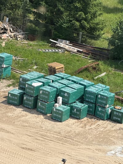 The image, taken from an aerial perspective in Mikołajki, Poland, shows a construction site or storage area on a sunny day. The primary focus is a large stack of green-wrapped building materials, likely blocks or bricks, organized neatly on the ground. Each package displays the "H+H" logo prominently. The surrounding area includes lush green grass and trees, suggesting an outdoor, possibly rural or semi-rural, setting. In the background, there are piles of lumber and metal, along with fencing and a ladder, indicating ongoing construction or demolition work. The ground is a mix of dirt and sand with visible tire tracks, reinforcing the idea of a work-in-progress environment. There are no people visible in the image, and the overall impression is one of a functional, outdoor workspace.