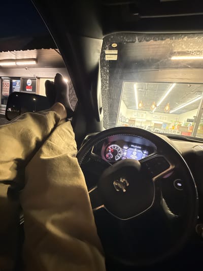 This image captures a person's perspective from inside a vehicle at night. The driver's hands, wearing dark socks, are resting on their lap, which is covered by a tan-colored garment, possibly pants or a blanket. The steering wheel of a modern vehicle, identified by the "RAM" logo on the dashboard display and the prominent "RAM" emblem on the steering wheel, is visible in the foreground. The dashboard lights and the reflection of overhead lighting suggest it's nighttime.
Through the windshield, a brightly lit establishment is visible, likely a laundromat or a similar service-oriented business. Signs advertising "20 WASHERS" and "60 WASHERS" are prominent, along with what appears to be a "REFER A FRIEND $50" promotion from "ATAX." The exterior lights create a slightly hazy or foggy atmosphere, common in such settings. The overall scene suggests the driver is waiting, perhaps for laundry to finish or for some service at the establishment. The lighting and the parked car suggest a moment of pause or waiting.
