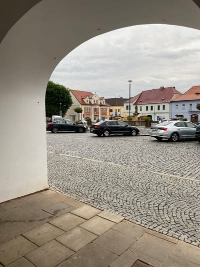 The image depicts a view through a large white archway onto a cobblestone plaza in Bílina, Czechia. Several cars are parked on the plaza, including a silver sedan and two dark sedans. Buildings with red tiled roofs line the plaza, some featuring ornate facades and windows. A street lamp is visible in the distance. The sky is overcast with clouds. The foreground shows large square paving stones leading up to the archway.
