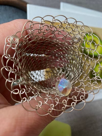 A close-up, top-down shot shows a person's hand holding a decorative, silver wire-mesh cylindrical object. The object is open at both ends and has an intricate, looped pattern. Inside the cylinder, near the bottom, a faceted, iridescent bead is visible. The background is blurred but suggests an indoor setting with what appear to be slices of avocado in the upper right quadrant. No specific location in Bílina, Czechia can be determined from the visual information provided.