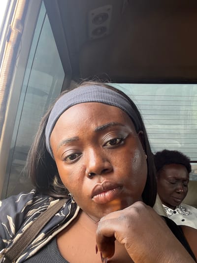 In Kugbo, Nigeria, a dark-skinned woman is photographed from the chest up, leaning on her hand. She is wearing a grey headband and a black patterned top. Her eyes are partially closed and she has a nose ring. Behind her, another woman is visible with her eyes closed, wearing a white shirt with a black collar and a patterned detail. The background appears to be the interior of a vehicle, with a window on the left and a dark ceiling with speakers visible at the top.
