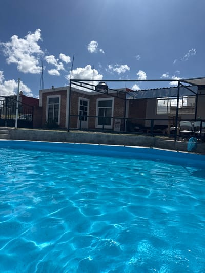 The image depicts a bright, sunny day with a clear blue sky dotted with white clouds. In the foreground, a bright blue swimming pool dominates the lower half of the frame, with sunlight reflecting and creating rippling patterns on the water's surface.
Beyond the pool, a low-lying, brick-faced building is visible. This structure has several windows and doors, and a black metal framework extends over part of it, supporting a translucent canopy. There are chairs and tables visible under this covered area, suggesting an outdoor seating or entertainment space. A black metal fence runs along the edge of the property, separating it from a sloping yard and a street where a vehicle can be partially seen. The architecture and the presence of a water tank on the roof of the building suggest a residential or small commercial establishment. The overall setting appears to be in a sunny, possibly rural or semi-urban, environment. The location is identified as San Baltazar Torija, Mexico.