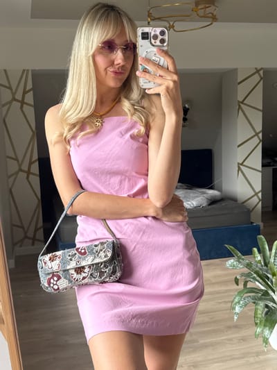 A blonde woman is standing in a bedroom, taking a mirror selfie. She is wearing a pink dress, pink sunglasses, and a gold necklace with a star pendant. She is holding a beaded handbag in her left hand. The bedroom has a blue bed with grey bedding, and a large mirror on the left side of the frame. There is a plant in a white pot on the right side of the frame. The wall behind the woman has a geometric gold pattern. The image is taken indoors during the day.
