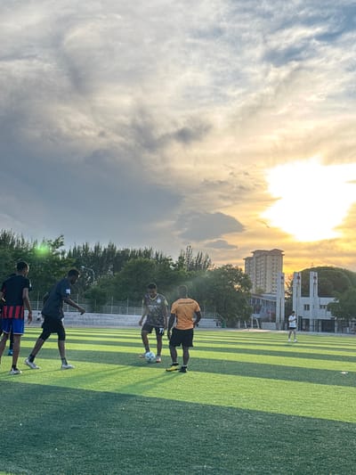 The image depicts an outdoor artificial turf soccer field in Linghe, China, during what appears to be late afternoon or early evening, as indicated by the warm, low-angle sunlight and dramatic cloud formations. Five young men are actively engaged in a soccer game.
In the foreground, four individuals are spread across the field. The person on the far left wears a black shirt with red and blue stripes and blue shorts. Next to him, another individual in a dark grey shirt with the number "95" on it and black shorts is in motion, appearing to control the ball. In the center of the action, a player in a patterned black and green jersey with the number "77" is kicking a soccer ball. To the right of him, a person in an orange shirt with "McLAREN" visible and black shorts stands near the ball.
Further in the background, a fifth individual, wearing a white shirt and black shorts, walks across the field away from the main group.
The environment includes a well-maintained green artificial turf field with distinct stripe patterns. In the mid-ground, there is a low fence, trees, and a street lamp. Beyond the trees, city buildings are visible, including a tall residential building and a white, modern-looking structure with arched entrances, suggesting an urban or semi-urban setting within Linghe. The sky is a prominent feature, filled with dramatic, sunlit clouds, with the sun itself obscured but its bright light casting long shadows across the field.