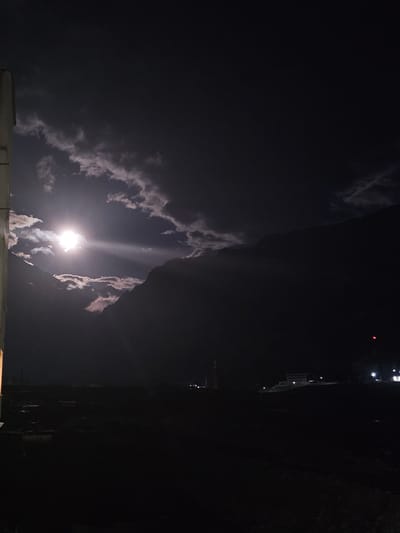 The image shows a nighttime scene in the city of Tyrnyauz, Russia, under a moonlit sky. The moon is partially obscured by clouds, casting a bright glow through the atmospheric haze and illuminating the surrounding mountains. In the foreground, a dark silhouette of a building or structure is visible on the left. The middle ground displays a dark landscape with faint outlines of buildings and possibly some vehicles. In the distance, on the right side of the image, several illuminated buildings and a tower with a red light are visible. The overall impression is one of a quiet, dimly lit urban or semi-urban environment nestled within a mountainous region at night.