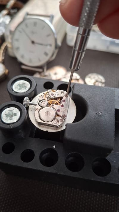 This media file depicts a close-up view of a watchmaker's hands working on a watch movement. The primary subjects are a person's fingers holding a watchmaking tool, and the intricate mechanics of a watch movement. The watch movement is silver-toned with small, gold-colored gears and ruby accents. It is held in place by a black watch repair stand with circular indentations. A portion of a white watch face with Roman numerals is visible to the left, suggesting the watch is in the process of being repaired or assembled. The setting appears to be a workspace, possibly a workshop, with a textured dark surface in the background. The lighting suggests it is daytime. There is no visible text in the image.