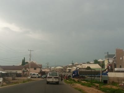 The image shows a street scene in Kabong, Nigeria, under an overcast sky. A paved road runs through the center of the frame, with a silver minivan driving away from the viewer in the foreground. To the left of the road, a row of buildings includes what appears to be a small shop or market stalls with corrugated metal roofs. Further back, a light brown building with a brown roof is visible, along with a white van parked in front. On the right side of the road, more buildings line the street, including a multi-story structure with a sign that reads "DOS STORES." A blue truck is parked on the right side of the road, and several people are walking and standing near the buildings. The environment appears to be a bustling urban or semi-urban area, with a mix of residential and commercial structures. Utility poles and power lines are visible throughout the scene. The overall impression is a typical day in a Nigerian town.