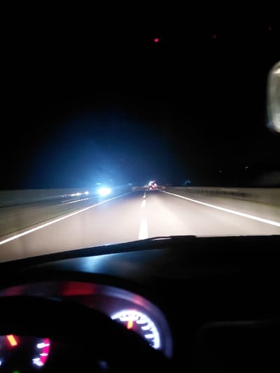 A nighttime view from inside a vehicle driving on a highway. The road stretches forward, illuminated by headlights. The highway appears elevated, with barriers visible on either side. The dashboard and steering wheel are partially visible in the foreground. The speedometer is illuminated. The overall scene is dark, with contrasting bright lights from the road and dashboard.
