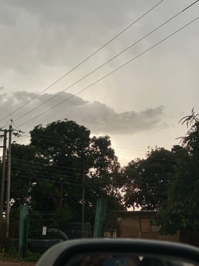 The image shows an outdoor scene in Maigizo, Nigeria, under an overcast sky with thick clouds. Power lines stretch across the sky, running from the lower left to the upper right. A utility pole is visible at the bottom. Trees with dense foliage are situated on the right, partially obscuring a building with a window and a fence. Part of a vehicle is visible on the right edge of the frame.