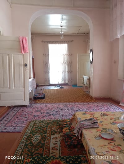 The image depicts the interior of a room in Beruniy, Uzbekistan. The room appears to be a living area or hallway, with multiple rugs covering the wooden floor. A large archway frames the central part of the room, leading to a doorway on the right.

In the foreground, a patterned rug with green and orange motifs is visible, partially obscured by a table covered with a floral-patterned tablecloth. A stack of folded fabric and a small red-trimmed dish sit on the tablecloth.

Beyond this, a larger pink and blue patterned rug stretches across the floor. A yellow rug is laid over part of the pink rug.

To the left, a white wooden door with decorative paneling is prominent. A pink cloth is draped over its top edge.

In the background, a large window with sheer curtains is the primary light source, illuminating the room. Several pairs of sandals are placed near the window. A white door is visible through the archway on the right, and a small table with a patterned cloth stands near it. A dark, oval-shaped mirror hangs on the wall to the right of the doorway. The walls are painted a light pink color. The ceiling is visible through the archway and appears to be textured.