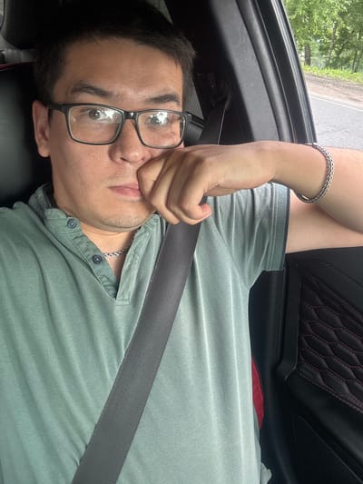 A medium shot captures a man sitting in the driver's seat of a car, with his right hand resting on his chin and his left arm on the car's door. He is wearing black-rimmed glasses, a light green collared shirt with two buttons, and a silver necklace. A seatbelt is visible across his chest. The car interior features black upholstery with red stitching on the door panel. Outside the car window, trees and a road can be seen, suggesting a rural or suburban environment. The lighting indicates it is daytime. The image was taken in Novoe Devyatkino, Russia.