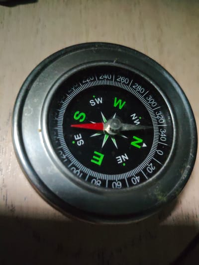 A close-up, overhead shot shows a black and silver compass with a metallic bezel, resting on a light-colored, textured surface, possibly wood. The compass face is marked with cardinal directions (N, S, E, W, NE, SE, SW, NW) and degree markings from 0 to 360. A red needle, indicating direction, points towards the 'S' marking. The image is slightly blurry and dark, with a narrow beam of light illuminating the center of the compass. There are no people, actions, or interactions visible in the image. The location of Naberezhnye Chelny, Russia is not determinable from this image.