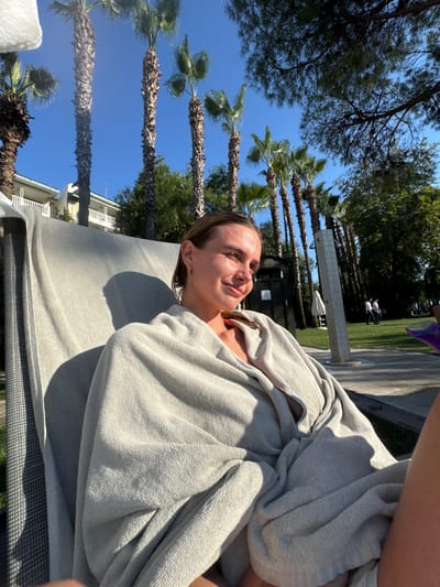 A young woman is reclining on a lounge chair outdoors, wrapped in a light gray towel. She is looking to her right, with her eyes partially closed and a slight smile on her face, suggesting relaxation. Palm trees and lush green trees are visible in the background, along with a building and a shower fixture. The scene appears to be a resort or hotel setting in Manavgat, Turkey, likely during a sunny day, as indicated by the strong sunlight and shadows.