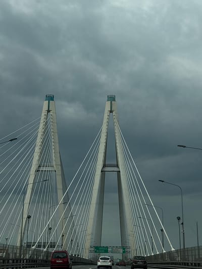 This is a photograph taken from a moving vehicle on a cable-stayed bridge, likely the Western High-Speed Diameter in or near Novosaratovka, Russia, given the signage visible. The sky is overcast with dark, dramatic clouds. Two massive concrete pylons of the bridge dominate the center and left of the frame, with numerous white cables extending from them to support the bridge deck. Several vehicles are visible on the roadway, including a red minivan in the foreground and a white car and a dark SUV further ahead. Streetlights are mounted along the sides of the bridge. Green directional signs are also present. The scene conveys a sense of imposing infrastructure against a moody sky.