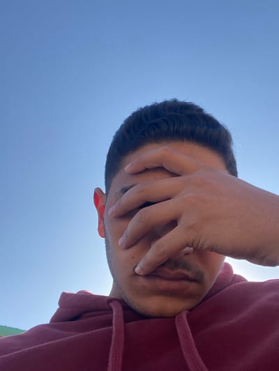 A man in a maroon hoodie shields his eyes from the sun. His hand covers much of his face, obscuring details except for part of his ear and mouth. He has dark hair with subtle waves. The background is a bright blue sky, indicating a sunny day. The perspective is from below, looking up at the man.