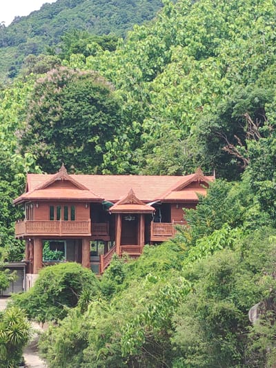 A traditional wooden house with a terracotta tiled roof sits nestled amongst lush green foliage and trees on a hillside. The house features intricate wooden carvings and balconies. The surrounding environment is dense with vegetation, suggesting a tropical or subtropical climate, possibly in a rural or natural setting. The image is taken from a distance, with the house partially obscured by trees. The sky is not visible. The location is identified as Baan Chaweng Noi, Thailand. There are no people or animals visible in the image, and no discernible actions or interactions are taking place.