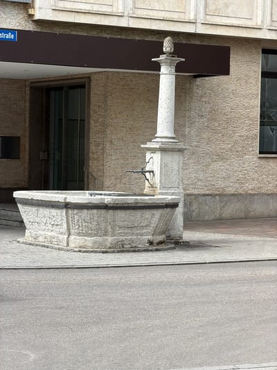 A stone fountain with a cylindrical pillar topped by a pinecone-like ornament stands on a paved area in front of a building. A blue street sign reading "lchstraße" is visible on the left side of the image. The building has a textured brick facade and large glass doors. The ground in the foreground is asphalt.