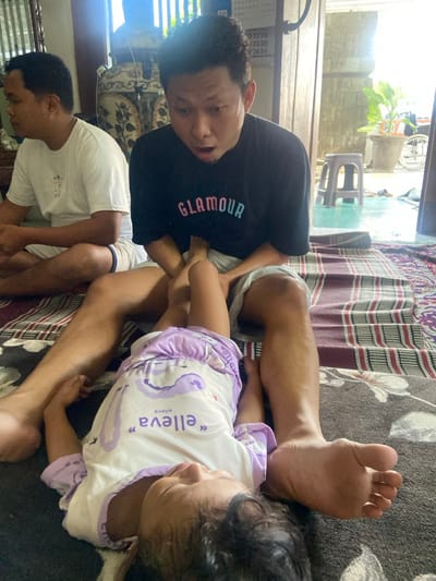 Three individuals are present in a domestic interior in Mojokerto, Indonesia. In the foreground, an adult male with dark hair, wearing a black t-shirt with "GLAMOUR" in pink and blue, sits on a dark grey rug. He displays an open-mouthed, focused expression while looking down at a child. The child, lying supine between his legs, wears a white and purple romper with "elleva" text and an elephant/whale motif. Her feet are elevated and positioned near the adult's midsection, with his hands on her lower legs. To the left background, a second adult male in a white t-shirt and light shorts sits, facing left, partially obscured. The interior space features patterned rugs (dark grey, and red/white striped) and an open doorway leading to an exterior area with light-colored tiles, a pale blue plastic stool, a potted plant, and a partial view of a wheeled apparatus. A calendar is mounted on a background wall.