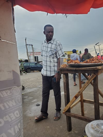 A man in a plaid shirt and dark pants stands in front of a food stall in Gwong, Nigeria. The food stall, made of rough-hewn wood, is laden with a large quantity of fried chicken. Other people are visible in the background, some attending the stall and others walking by. A blue van is parked on the side of the road. The scene appears to be outdoors during the day, with a bright, overcast sky. There are utility poles with wires visible in the distance. Some text is visible on a sign behind the food stall, reading "EXCEL DIAGNOSTIC CLINICAL SERVICE." Another sign with text is visible on a cardboard box in the foreground.