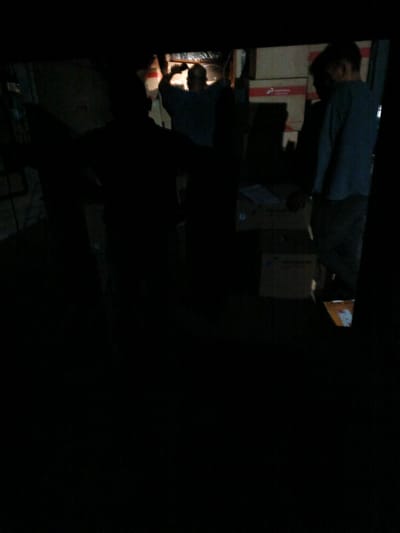 The image depicts a dimly lit interior space, likely a warehouse or storage area, with several individuals present. In the center, a person is holding up what appears to be a piece of equipment or a tool, possibly working on overhead infrastructure. To the right, another person stands facing away from the camera, wearing a blue shirt and dark pants, holding a box. A third individual, in silhouette, is positioned to the left, also holding an object. The background shows stacks of cardboard boxes, some with red labeling that suggests they are manufactured goods. The overall lighting is low, creating strong shadows and making it difficult to discern fine details. The scene appears to be a snapshot of work activity in progress. The location in Bireuen, Indonesia, cannot be determined from the visual information alone.