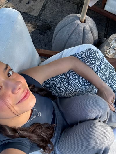 The image displays a female individual reclining outdoors in Huasca de Ocampo, Mexico, viewed from a high-angle, slightly off-center perspective. The subject is positioned with her head towards the upper left, looking directly at the camera, with natural sunlight illuminating her face. She has dark hair styled in braids visible over her left shoulder. She wears a dark t-shirt, gray ribbed pants, and a dark fabric with a white geometric pattern draped over her lower torso. Her left hand, adorned with rings on the index and ring fingers and dark nail polish, rests on her lap. A thin silver necklace with a circular pendant is visible around her neck. She is seated in a white upholstered chair with visible wooden elements. The ground is a dark gray flagstone or cobblestone patio. In the background, to the right, a large, fluted, pumpkin-shaped gray stone umbrella base holds a light-colored wooden pole. Further right, on a stone surface, two clear glasses are visible. The scene is brightly lit by natural sunlight, casting distinct shadows.