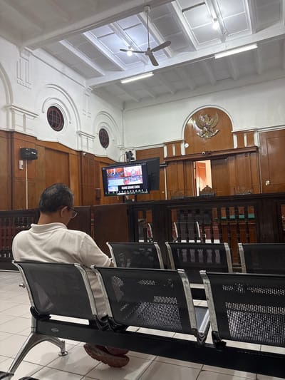 A man sits on metal chairs in a courtroom. A screen displays a virtual meeting. Wood paneling lines the walls, featuring circular windows and an emblem of an eagle holding a shield. Overhead, a white ceiling with fluorescent lights and a fan is visible. The chairs appear to be lined up in rows. The courtroom looks formal, with classic architectural details.