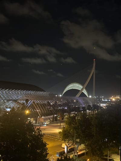 The image captures a nighttime view of the City of Arts and Sciences in Valencia, Spain. The architectural complex is illuminated, with the striking cable-stayed bridge dominating the scene. The bridge's white pylon, topped with a red light, stands tall against the dark, cloudy sky. The distinctive ribbed structure of the L'Hemisfèric is visible in the background, with its arched roof glowing with interior lights. In the foreground, dense trees with illuminated elements suggest parkland or pedestrian areas within the complex. A street with passing vehicles, including a white van, is visible below the structures, with streetlights casting a warm glow on the asphalt. The overall impression is one of modern, illuminated architecture set against a nocturnal urban landscape.
