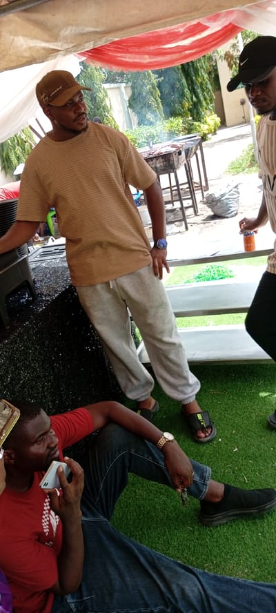 An outdoor gathering is taking place under a tent with a red and white canopy. In the foreground, a man wearing a red t-shirt and blue jeans is sitting on artificial grass, holding a light blue smartphone to his ear. He is wearing a gold wristwatch on his left wrist. To his right, another man wearing a brown baseball cap and a horizontally striped brown t-shirt is standing. He has a watch on his left wrist. Between him and another person to his right, there is a barbecue grill with food on it, emitting smoke. The person to the right is wearing a black baseball cap and a horizontally striped t-shirt, and is holding an orange bottle. The background features lush green trees and buildings. The scene appears to be in Giginyu, Nigeria.