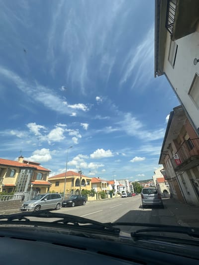 The image depicts a street scene in Macedo de Cavaleiros, Portugal, as viewed from inside a vehicle. Buildings line the street on both sides, with a mixture of architectural styles. The sky is visible above, filled with wispy clouds. Parked cars are visible along the side of the road, with a few other vehicles in the distance. The scene appears to be taken during the daytime, with bright sunlight and clear visibility. The car's interior elements, such as the windshield wipers, are also visible.