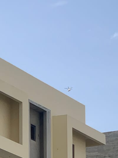 An outdoor scene captured under a clear, bright blue sky in Moba, Nigeria, depicts the upper portions of architectural structures. A prominent light-beige or light-yellow building, possibly under construction or recently completed, occupies the lower and left-mid sections of the frame. This building features sharp geometric lines, flat roof sections, and visible cut-outs or recesses, including a dark rectangular opening within a larger vertical recess on the left side. In the upper right quadrant of the sky, a distant helicopter with a white or light-colored body and visible rotor blades is in flight, presenting with faint red or orange markings. A rough-textured, grey wall or structure is partially visible on the far right, behind the main building. No individuals or direct interactions are visible.