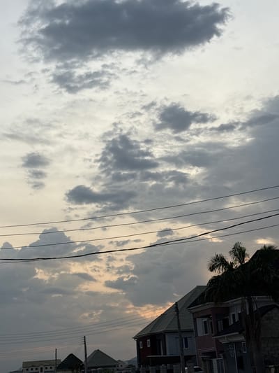 The image depicts a cloudy sky over a residential area in Angwan Sayawa, Nigeria. The sky is filled with numerous grey clouds, with some lighter patches suggesting the presence of the sun behind them. Several dark power lines stretch horizontally across the upper portion of the frame. In the lower part of the image, several houses with distinct architectural styles are visible, some featuring pitched roofs. Palm trees are also present, with one prominently silhouetted against the sky on the right side of the frame. The overall impression is of a somewhat overcast day in a Nigerian urban or suburban setting.