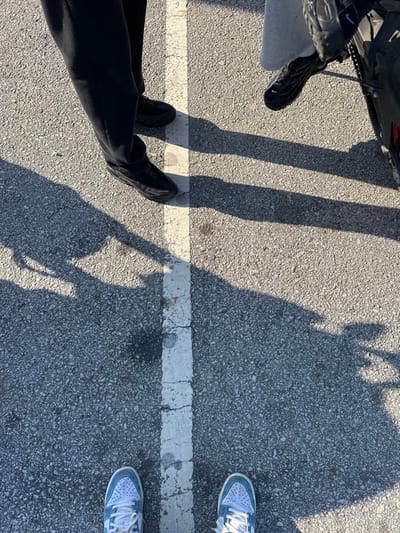 An overhead view depicts three individuals standing on an asphalt surface in Mirandela, Portugal. A worn white line bisects the frame vertically, positioned slightly left of center. To the left of the line, the lower legs of a person wearing dark pants and black shoes are visible. To the right of the line, the lower leg of a second person wearing light grey pants and black sneakers is present, positioned alongside the partial view of a black motorcycle's wheel and frame. At the bottom of the frame, the feet of a third individual, wearing light blue and white sneakers, are positioned on the asphalt, straddling the white line from the perspective of the camera holder. Multiple elongated shadows are cast across the textured grey asphalt surface, indicating a low sun angle.