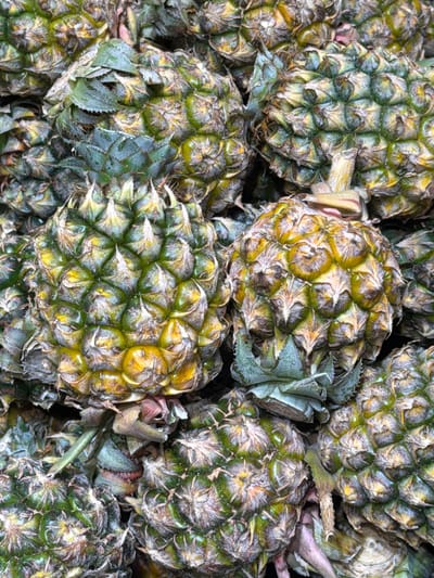 A close-up, overhead view showcases a pile of ripe pineapples, their textured green and yellow skins forming a dense mosaic. The pineapples, with their spiky crowns, are stacked closely together, obscuring much of the surrounding environment. The lighting is diffuse, highlighting the intricate patterns of the fruit. There are no people visible in the image. The scene appears to be a market stall or grocery display in Baan Chaweng, Thailand, where such tropical fruits are commonly found.
