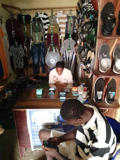 The image depicts a small, cluttered shop in Serti, Nigeria, likely a clothing and accessories store. Two young men are present. One, wearing a cream-colored garment with a diamond pattern, sits behind a wooden counter, engaged with a mobile device or a transaction device. On the counter, two small transaction devices, one green and one blue, are visible, along with various other small items. The second young man, wearing a black and white striped shirt, is seated in the foreground, also looking at a mobile phone.
The shop is densely stocked with clothing items, including shirts and jeans, hanging on display racks and chains. A fan is positioned behind the counter. Shelves on the right side of the image display an assortment of footwear, primarily black shoes and sandals. The overall impression is of a small, busy retail space in a local Nigerian setting.