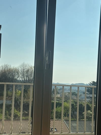 The image depicts an outward view from inside a building, likely through a glass window or balcony door, located in Guimarães, Portugal. A dark-colored vertical window mullion with a light-colored handle occupies the center-right of the frame. Beyond this, a light-colored metal balcony railing with multiple vertical bars extends horizontally across the lower and middle portions of the view.
The scene beyond the railing reveals a varied landscape under a clear, pale blue sky. In the mid-ground, numerous residential rooftops are visible, predominantly featuring reddish-brown terra cotta tiles, with some areas of darker roofing. Green foliage from trees and shrubs is interspersed among these structures and extends into the background. Some deciduous trees with bare branches are also present. The far distance shows a hilly terrain with additional scattered buildings and vegetation, indicating a suburban or semi-rural environment. No people or active interactions are visible. A faint smudge is present on the upper left pane of the glass.