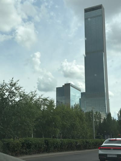 The image is an outdoor, eye-level, medium shot of a cityscape under a cloudy sky. The primary subjects are modern, glass-paneled skyscrapers with geometric designs. One skyscraper, in particular, is a tall, rectangular structure with a distinct horizontal banding pattern and a sleek, metallic top. Adjacent to it, another building with a similar glass facade and tiered construction is visible. The scene is framed by lush green trees and manicured hedges lining a paved road.

In the foreground, the rear of a white car is partially visible. The car has distinctive red LED taillights and a license plate with the text "011DDD08". This suggests a street-level perspective, possibly from another vehicle or a sidewalk. The sky is overcast with scattered clouds, indicating a potentially bright but not sunny day.

The overall impression is one of urban development and modern architecture within a well-maintained green environment. The location cues, such as the architecture style and the license plate format, combined with the provided location context of Astana, Kazakhstan, suggest this is a scene from a developed area within that city. No people are clearly visible, and the activity seems to be static, capturing a moment in the urban landscape rather than a specific event. The weather appears to be temperate, with no strong indications of extreme conditions. The image is captured in landscape orientation, with the buildings appearing on the left side of the frame and the road on the right.