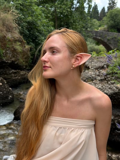 The image shows a close-up of a young woman with blonde hair and elf-like ears. She is wearing a light-colored, strapless dress. Her head is tilted down, and she is looking to the side with a gentle expression. The background is blurred, but appears to be a natural setting with greenery, rocks, and a hint of a stone bridge and water, possibly a stream or waterfall. The lighting suggests it is daytime, likely outdoors. The context provided indicates the location is Podgorica, Montenegro. There is no visible text in the image.