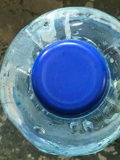 The image is a close-up, top-down view of a blue plastic bottle cap on a clear plastic water bottle. The bottle appears to be partially filled with water, as evidenced by condensation and a few small bubbles inside. The background is a dark, textured surface, possibly a table or countertop. The lighting is diffused, with some highlights reflecting off the plastic. There are no people or any other identifiable objects in the frame. The location is New Stadium, Nigeria.