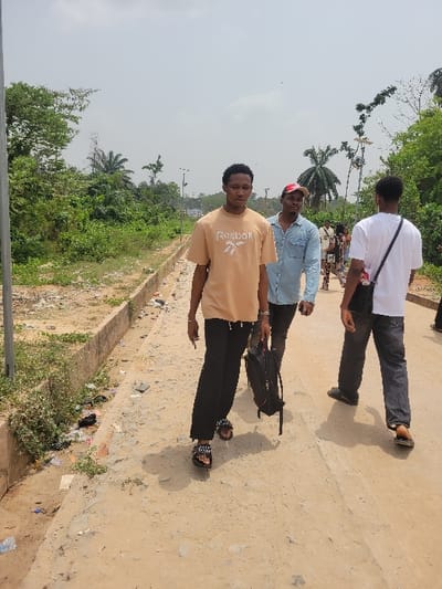 An outdoor scene in Candelaria, Colombia, depicts multiple individuals walking along an unpaved dirt path under an overcast sky.

In the foreground, slightly left of center, a young male is observed walking towards the background, facing slightly left of the camera. He wears a beige short-sleeved t-shirt with the "Reebok" logo, black trousers, and dark open-toed slide sandals with white embellishments. He carries a black backpack in his left hand.

Immediately behind him and to the right, a second young male walks, wearing a light blue denim shirt and a red baseball cap. To the far right, a third young male is visible from behind, dressed in a white t-shirt, dark trousers, and dark slide sandals, carrying a black shoulder bag. Additional figures are partially visible further in the background, suggesting a larger group.

The ground consists of a light brown, uneven dirt or gravel path, showing signs of use and small debris. To the left of the path, a concrete curb runs parallel to the direction of travel. Beyond the curb is an unkempt strip of ground with green weeds, exposed dirt, and scattered litter including plastic bottles and papers. Dense tropical vegetation, including palm trees and various other trees, forms a green wall bordering both sides of the path and extending into the background. A grey utility pole is visible on the far left. In the distance, additional utility poles and a potential communications tower can be seen amidst the foliage. The sky is a uniform light grey, indicative of overcast conditions.