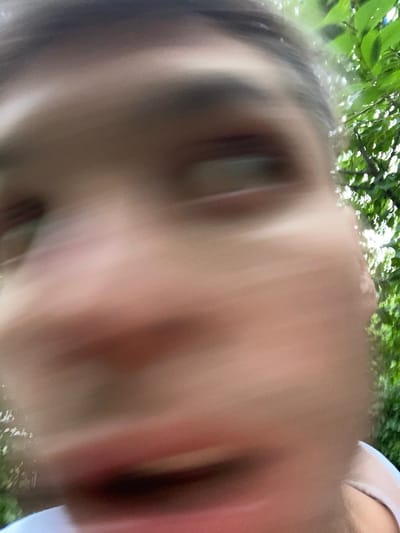 The image is a close-up, blurry photograph of a person's face, taken from a low angle looking upwards. The subject appears to be outdoors, with blurred green foliage and sunlight visible in the background, suggesting a daytime setting. The person's face is largely out of focus, with some features like an eye and cheekbone vaguely discernible. The light is bright, creating a somewhat washed-out effect across the image. There is no visible text. The overall impression is that of a candid, possibly accidental, shot taken in natural light.