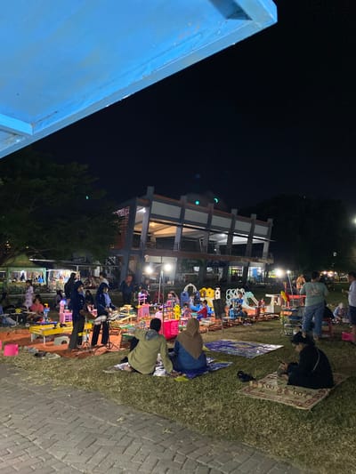 The image depicts an outdoor night market in Madiun, Indonesia, likely held in a public park or open space. The scene is illuminated by artificial lights, creating a warm and lively atmosphere. In the foreground, several people are seated on mats, engaged in various activities such as looking at their phones and interacting with each other. A woman in a black hijab is seated on a patterned rug, engrossed in her mobile phone. Next to her, a person in a green hoodie and blue shorts is sitting on a blanket, facing away from the camera.

The middle ground of the image is filled with various stalls and attractions typical of a night market. There are numerous colorful stalls displaying goods, and a play area with various toys and structures, including a small slide and a toy kitchen. Children and adults are seen browsing the stalls and playing in the designated area. In the background, a modern building with multiple levels stands out, its windows illuminated, suggesting it might be a community center or a commercial establishment. Trees surround the area, adding to the natural ambiance of the location. The overall impression is a vibrant and social gathering, characteristic of night markets in Indonesian cities.