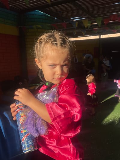 The image shows a young girl, possibly of European descent, with blonde braided hair and blue eyes, holding a can of "Rey Grito Fantasía" (King Shout Fantasy) foam spray. She is wearing a bright pink satin outfit and a black cape with colorful trim. Her expression is stern. In the background, other children and adults are present in what appears to be an outdoor event space with a covered ceiling adorned with colorful triangular flags. The ground is a dark green surface with scattered white specks, possibly confetti or foam. Sunlight casts strong shadows across the scene. The event is taking place at the "Parque Infantil Benjamín Vicuña Mackenna" located in Arica, Chile, a public park with recreational facilities.